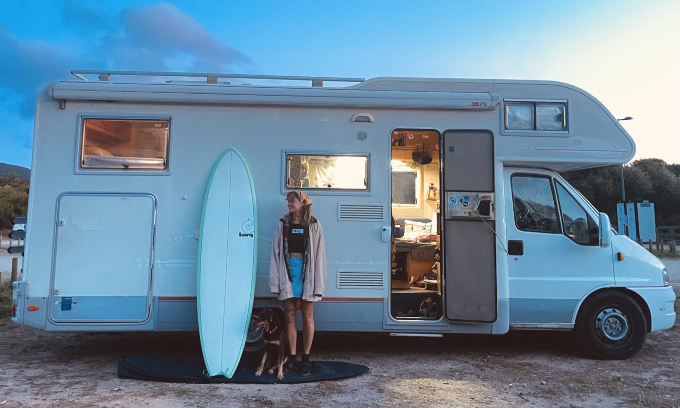 Karolina with her surfboard and dog beside the camper van at dusk